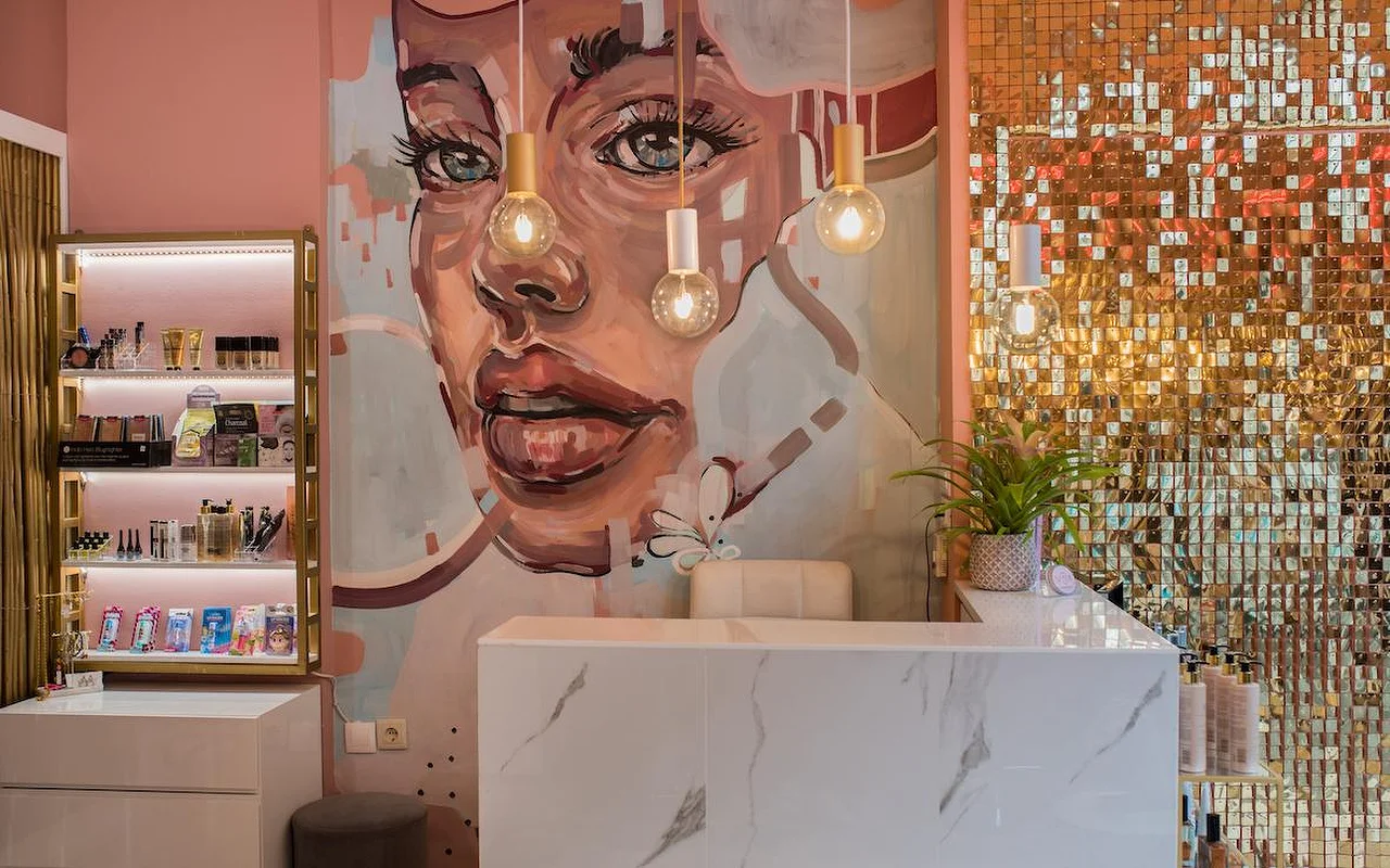 Un salone dalle pareti rosa con un grande murale dipinto del volto di una donna, una parete di paillettes dorate scintillanti, un bancone reception in marmo bianco e lampade a sospensione a bulbo scoperto.