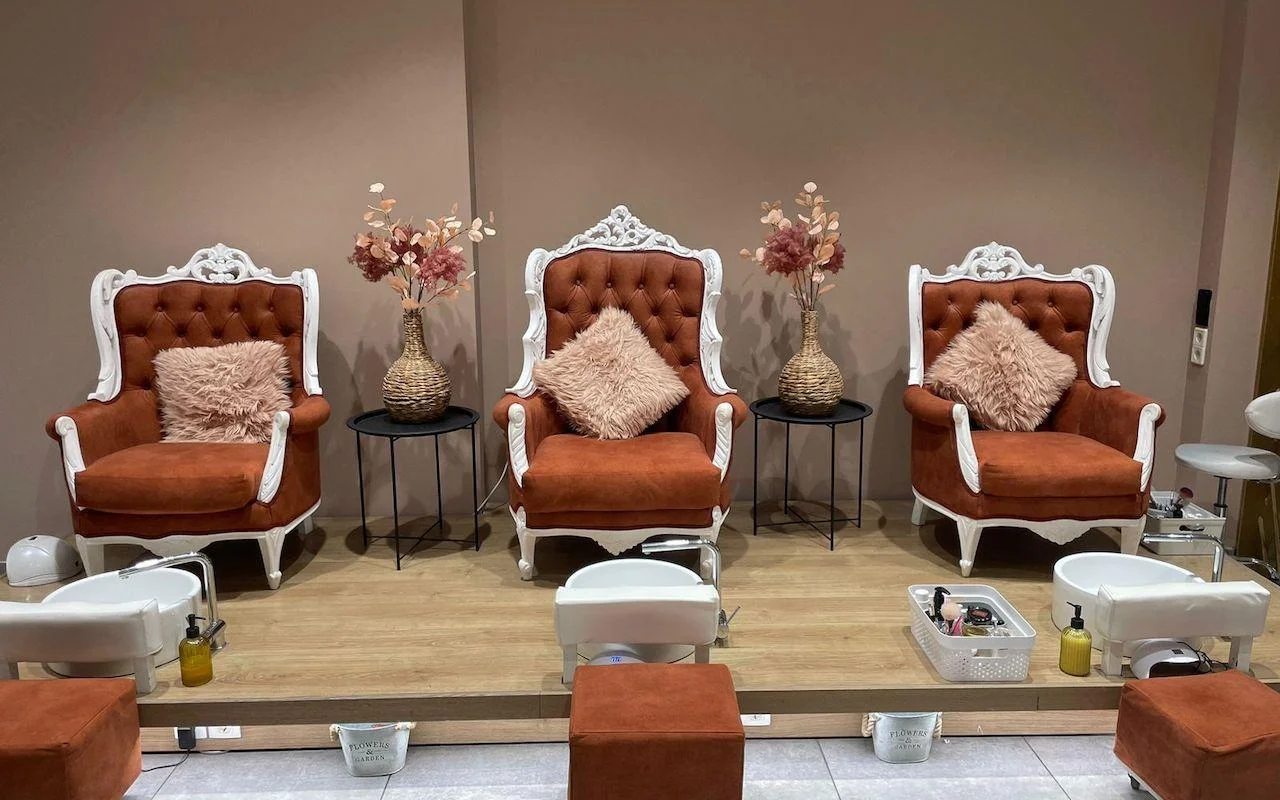 Tre poltrone eleganti con cornice bianca in velluto arancione bruciato, rialzate su una piattaforma di legno sopra vasche pedicure bianche, con fiori essiccati e cuscini rosa tenue tra i sedili.