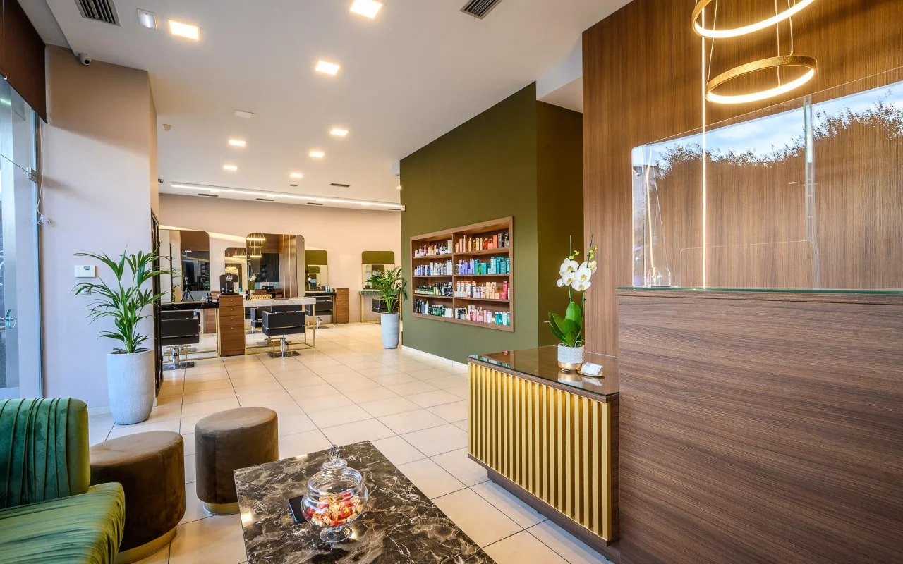 Un salone di parrucchiere di alta gamma con pannellature in legno caldo, una parete decorativa verde oliva con scaffali di prodotti, postazioni styling con grandi specchi, un bancone reception con listelli dorati e sedute in velluto verde sotto lampade a sospensione dorate circolari.