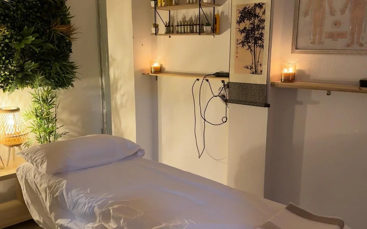 Una sala trattamenti dalla luce soffusa con un lettino da massaggio rifatto in lino bianco. Candele accese sono sistemate su mensole in legno accanto a un rotolo di inchiostro in bambù e a una tavola di agopuntura incorniciata, con una pianta rigogliosa e una lampada intrecciata luminosa nell'angolo.