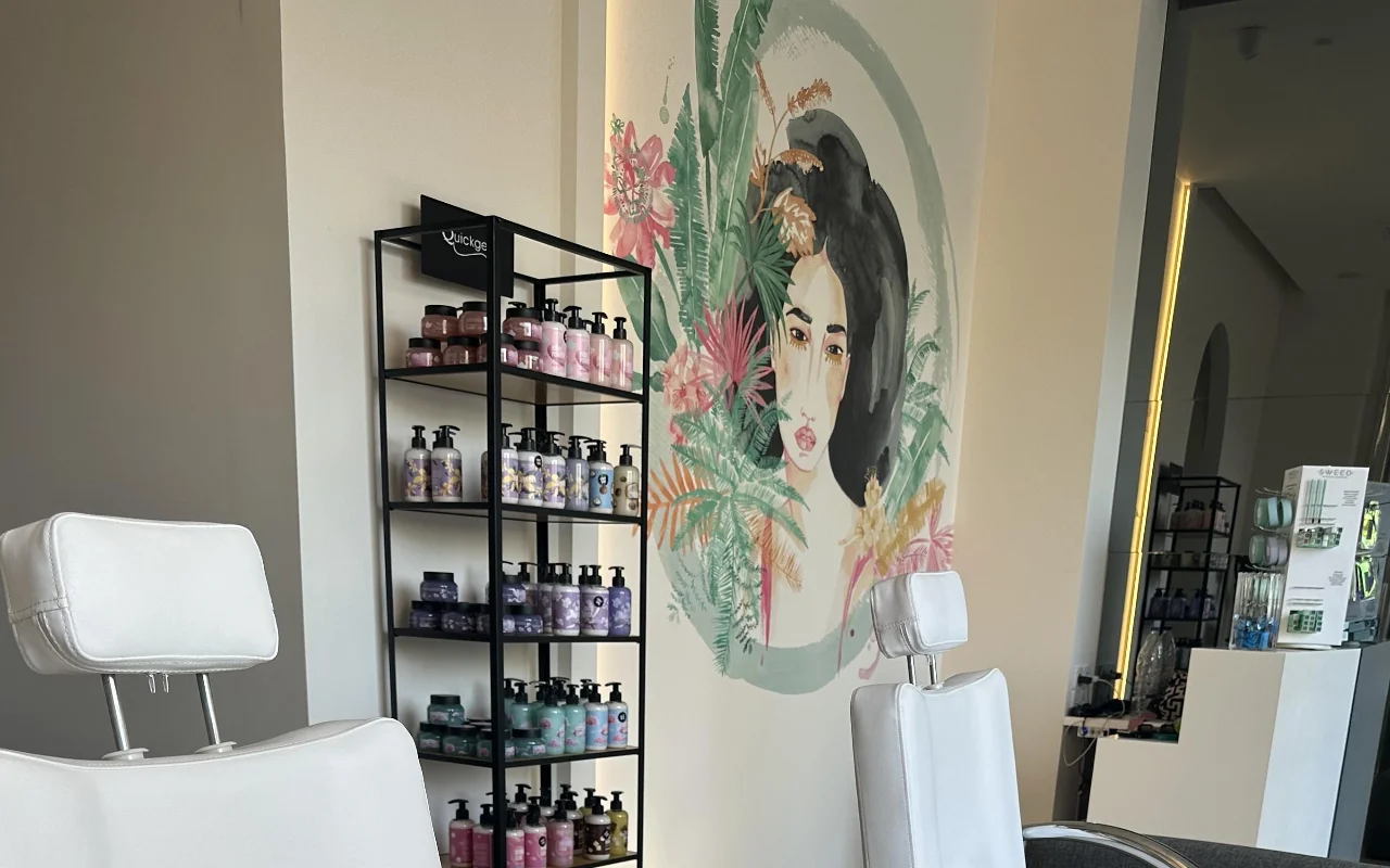 Un angolo studio con due poltrone da trattamento bianche reclinabili, uno scaffale nero di prodotti, uno specchio retroilluminato e un murale floreale del volto di una donna incorniciato da fogliame tropicale.
