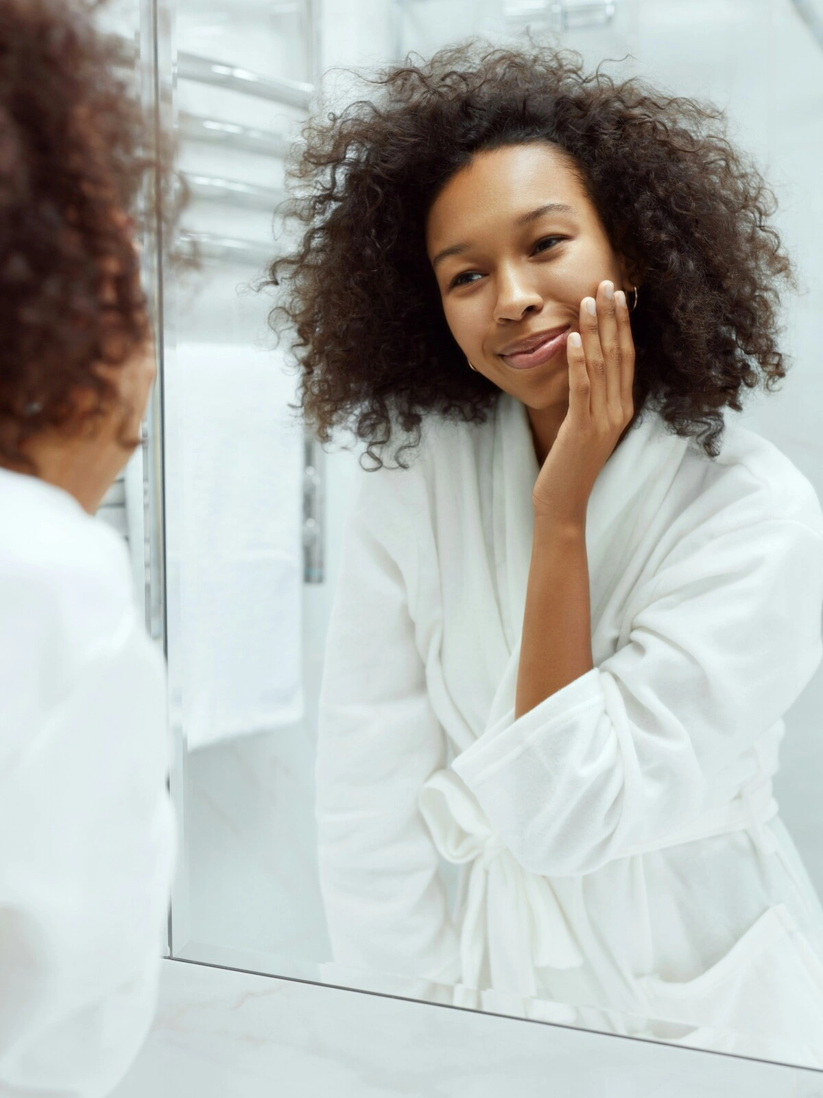 Una donna in accappatoio bianco è in piedi davanti a un grande specchio nel suo bagno pulito e bianco e sfiora delicatamente la pelle liscia del viso