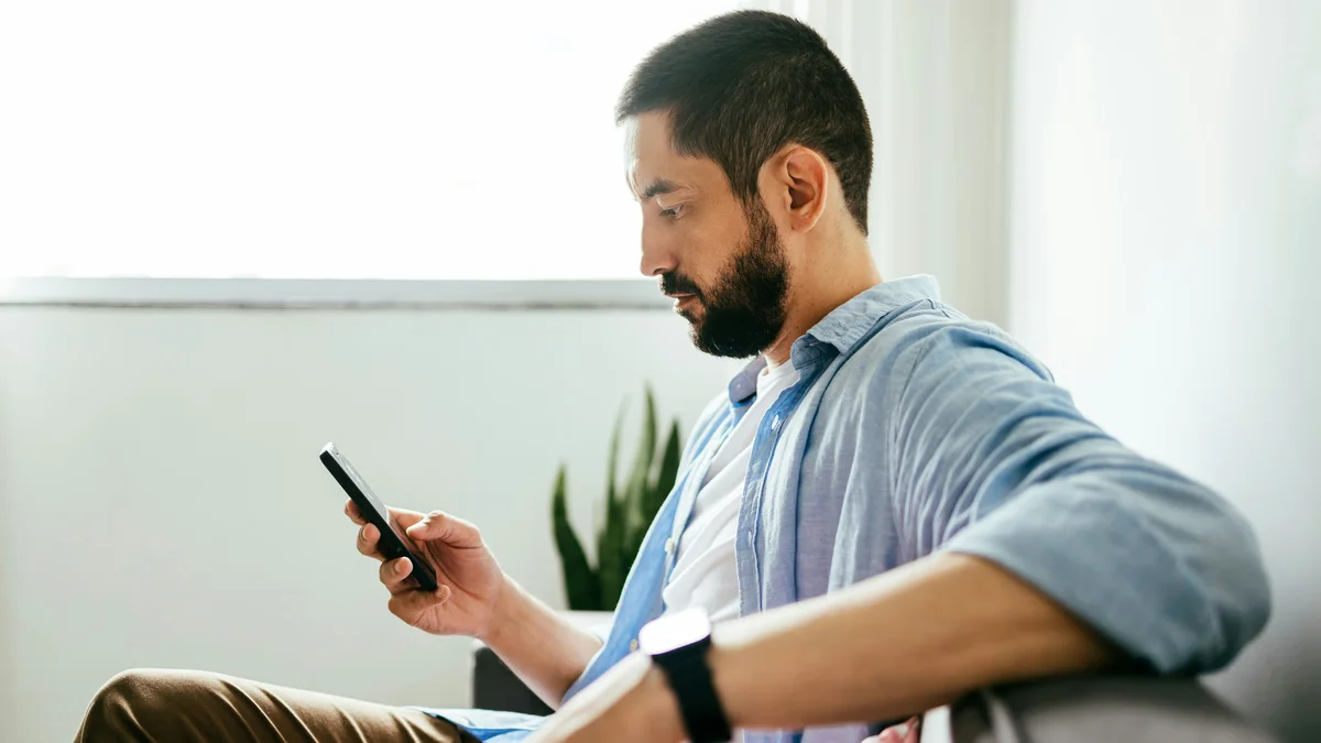 Vista laterale di un uomo seduto sul divano che appoggia un braccio sullo schienale e con l’altra mano prenota un trattamento sul telefono